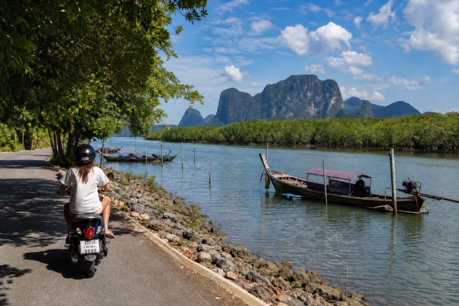 Helmeted scooter ride along the Krabi River with longtail boats on the water and mangroves nearby
